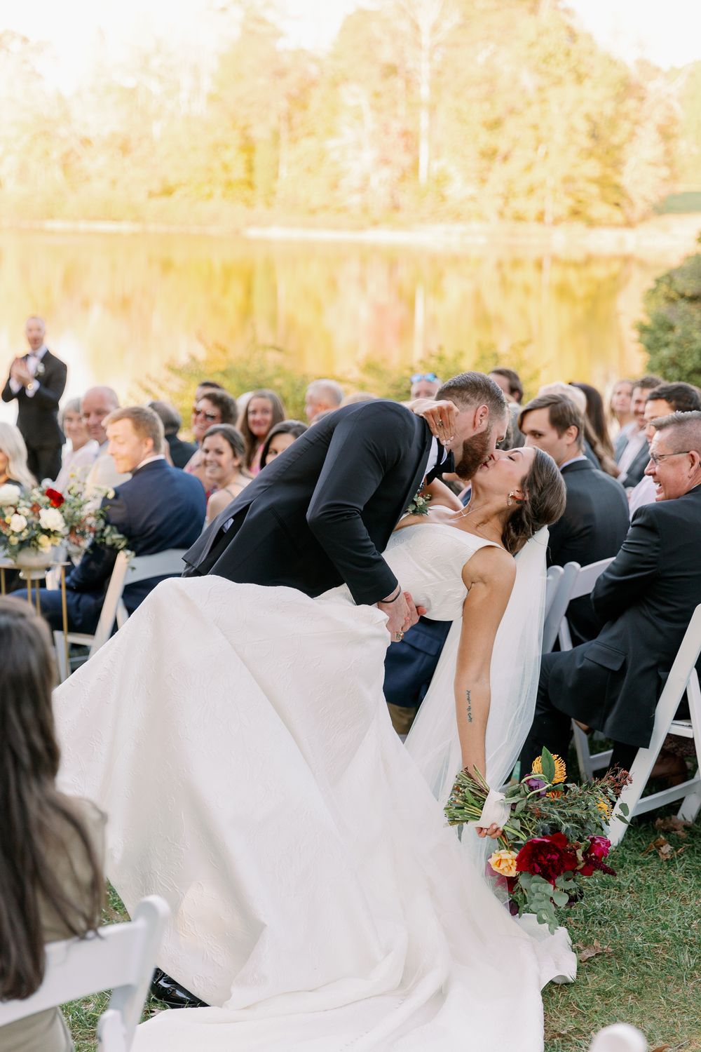 Wedding ceremony guests watch as couple shares romantic moment against autumn lakeside backdrop.