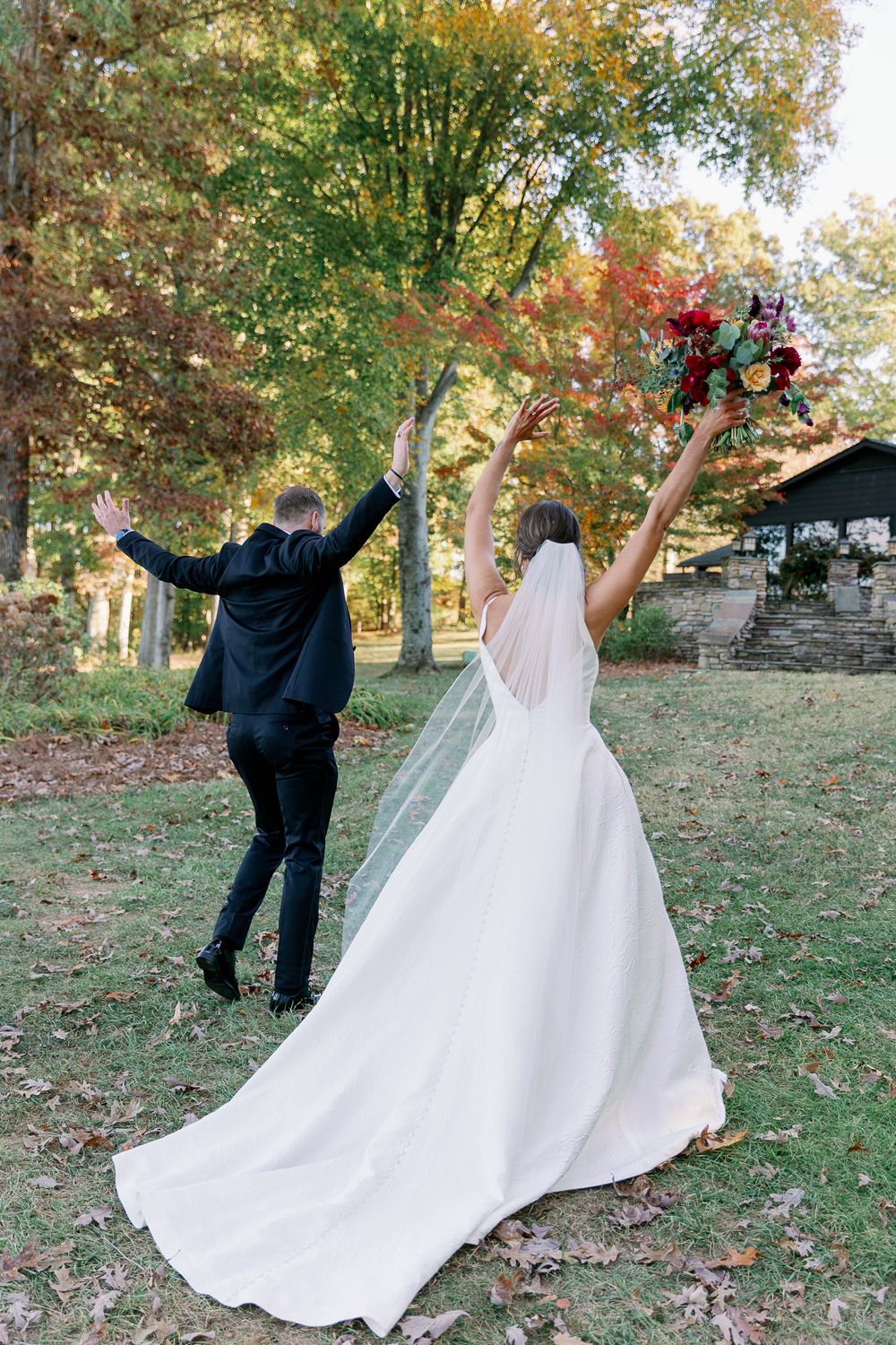 Newlyweds celebrate with joyful poses on grassy hill during autumn wedding photoshoot.