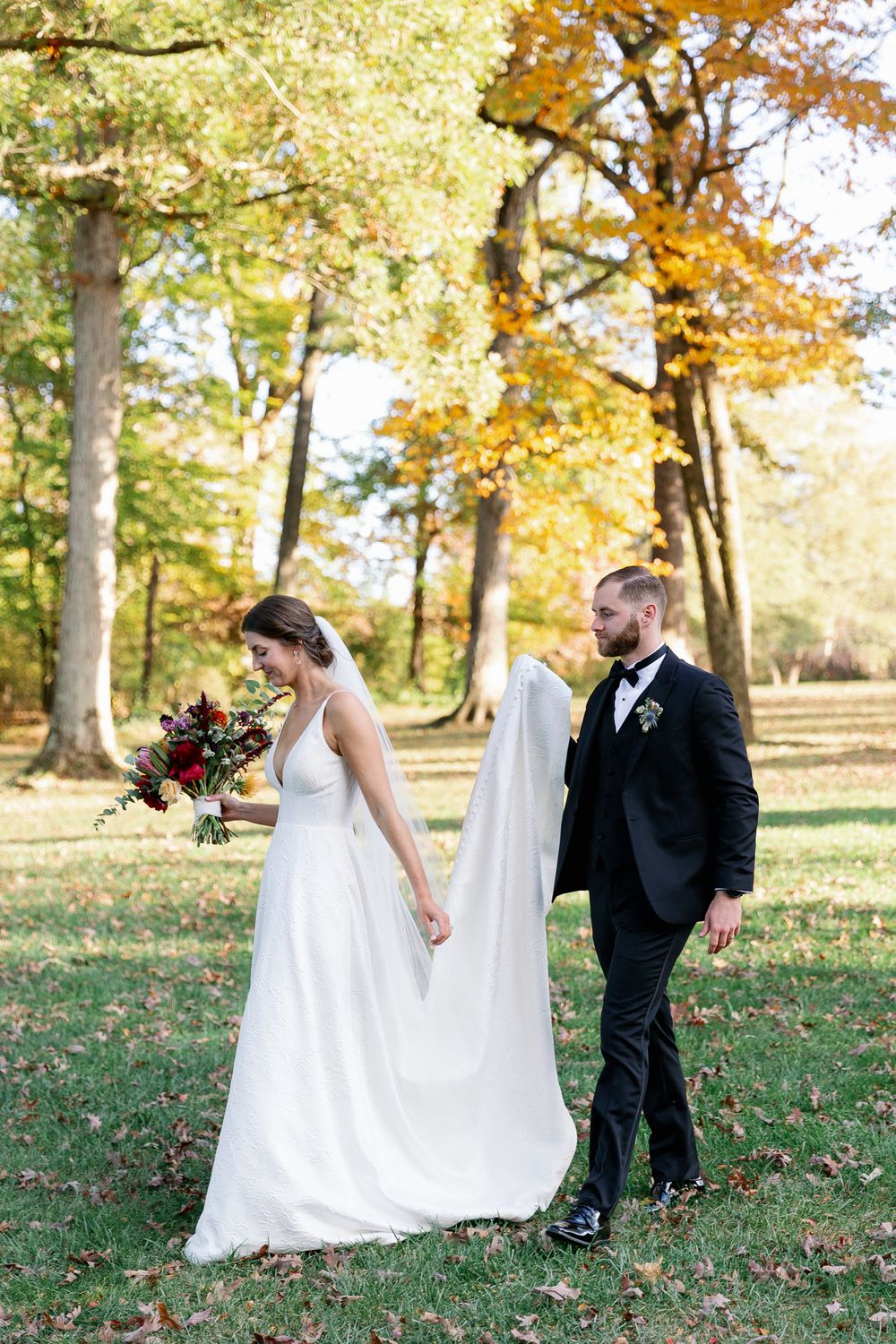 A couple in wedding attire walks through a park with golden autumn foliage.