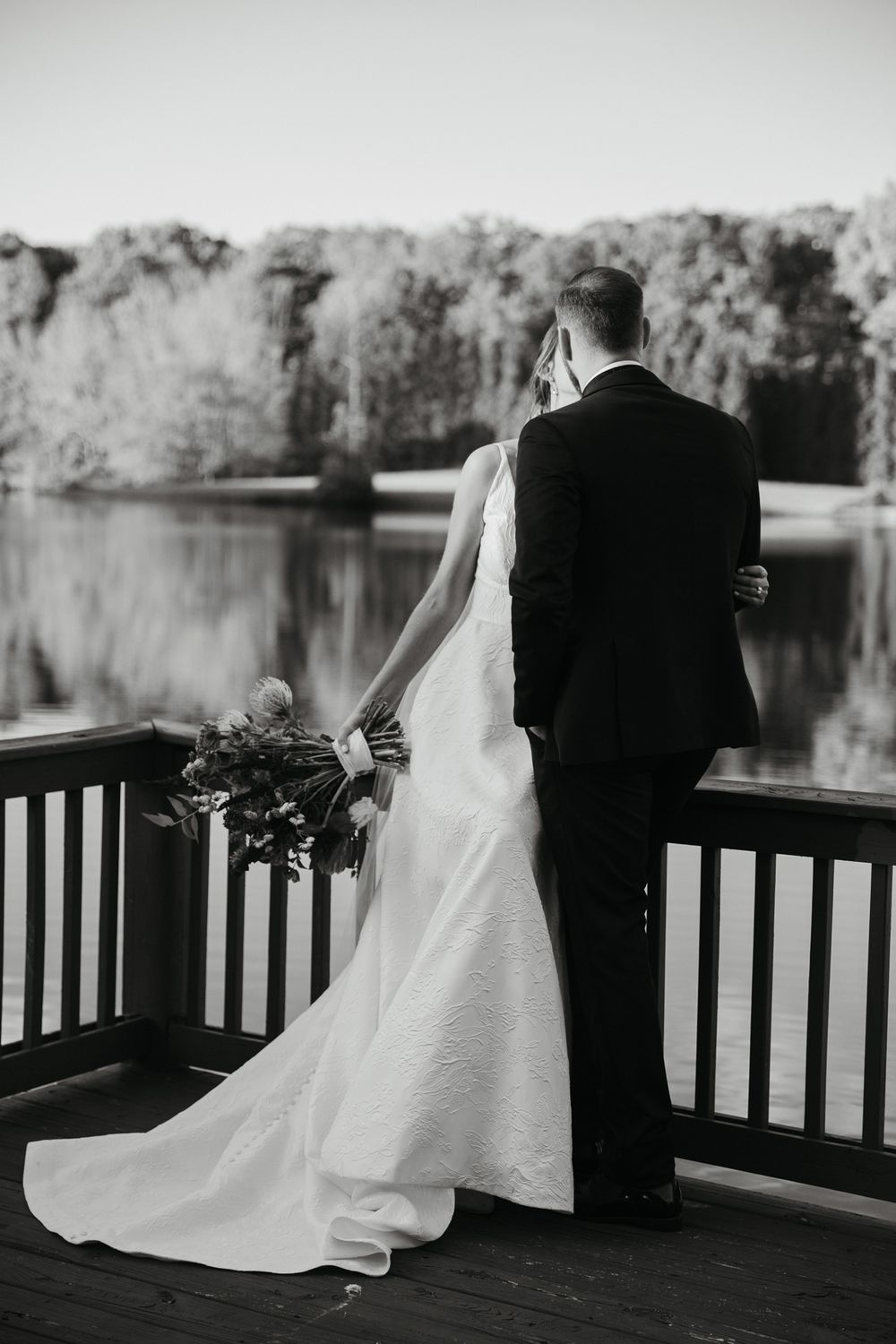 Newlyweds sit together on a wooden bench overlooking a serene lake.