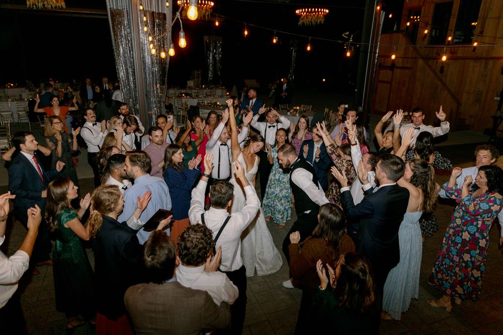 Lively wedding reception crowd dances under ambient lighting in rustic venue.