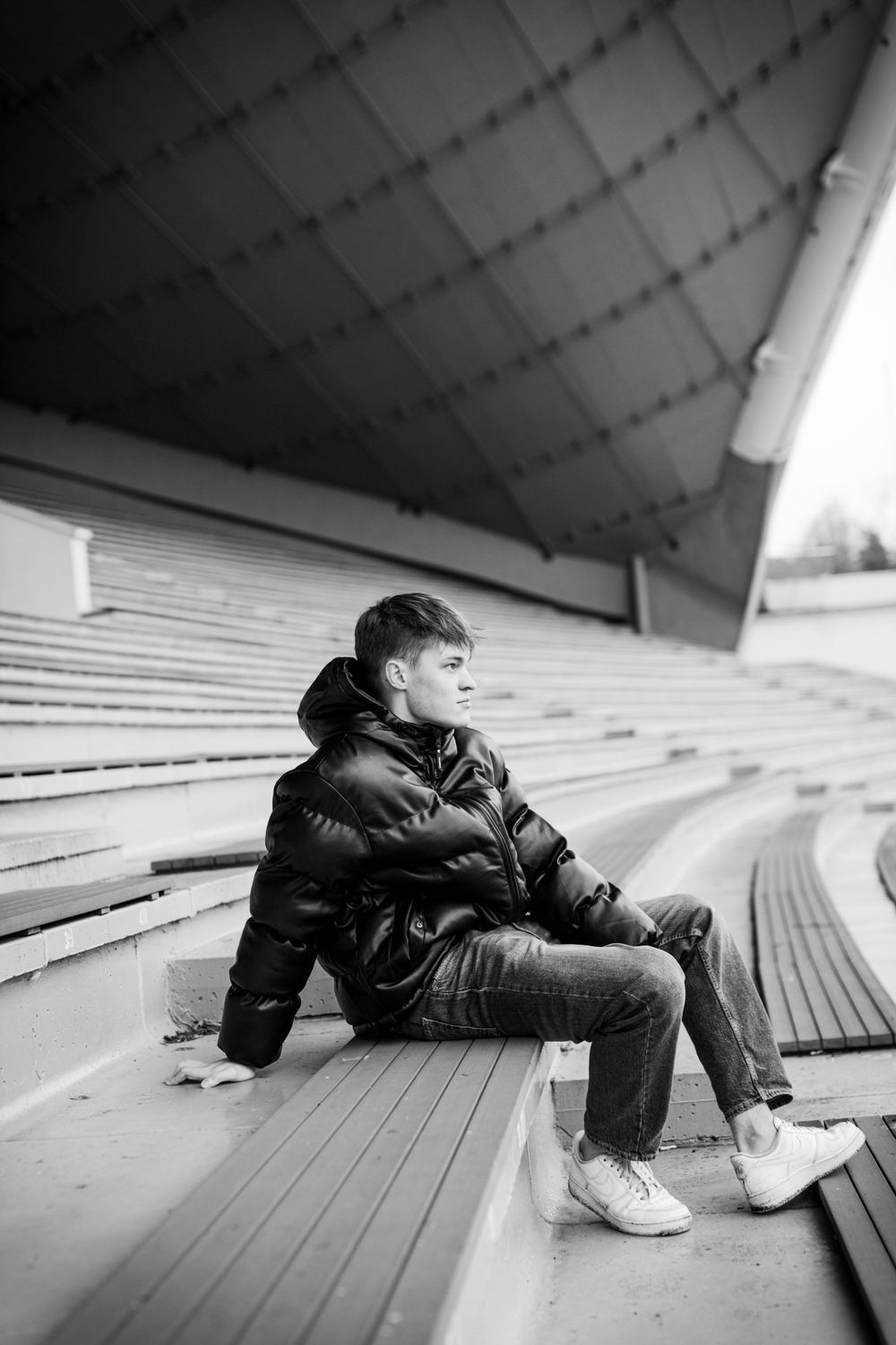 Black and white photo of person sitting on stadium bleachers wearing puffy jacket.