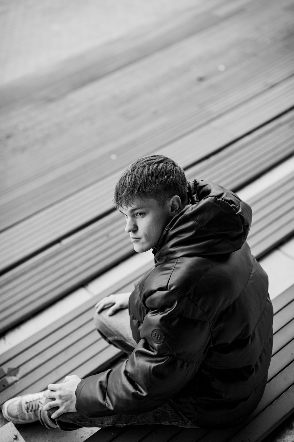 Monochrome portrait of person in dark jacket sitting on wooden bleachers.