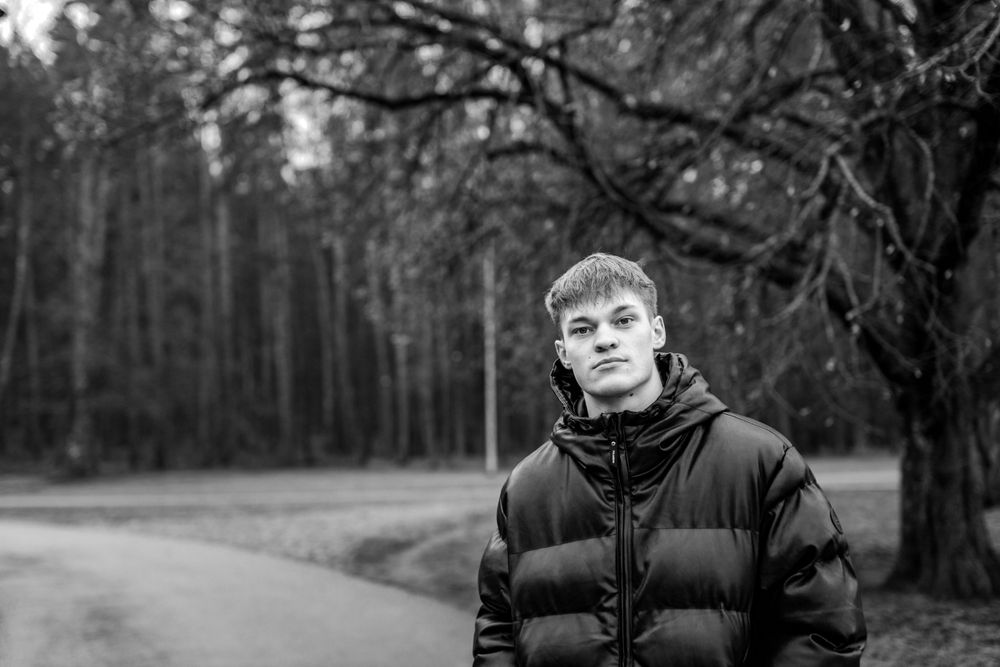 Person wearing puffer jacket stands on path in wooded area, black and white photo.