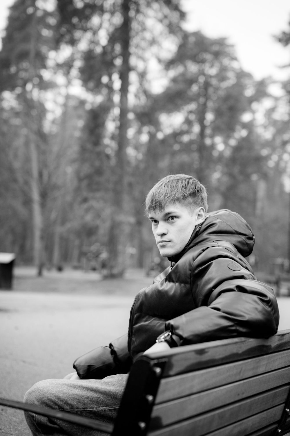 Person in black winter jacket sits on park bench in monochrome forest setting.