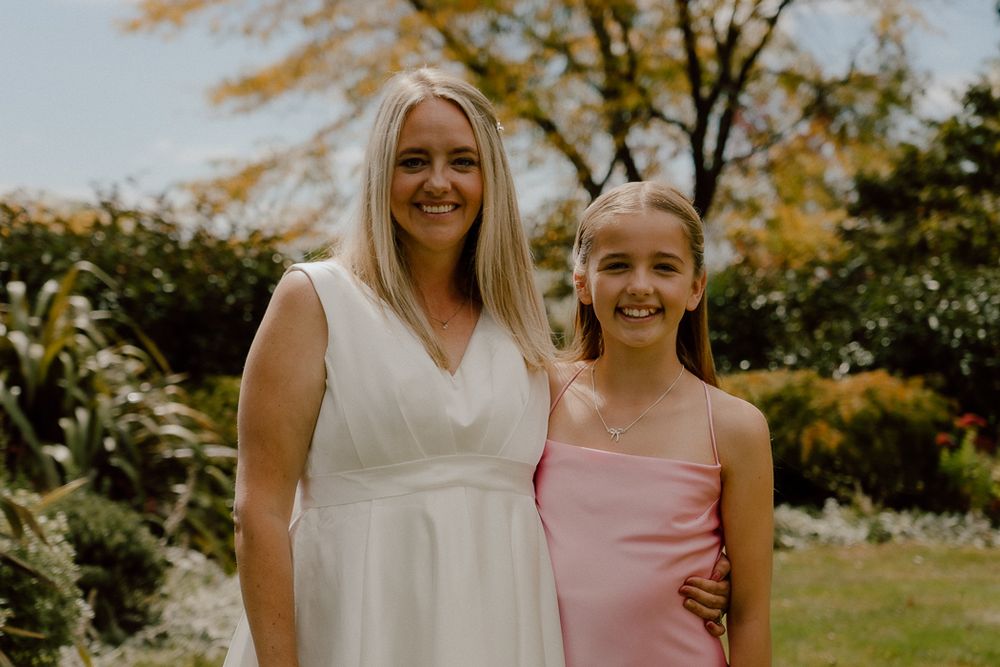 A woman in white dress and young girl in pink dress smiling together outdoors in a garden setting.