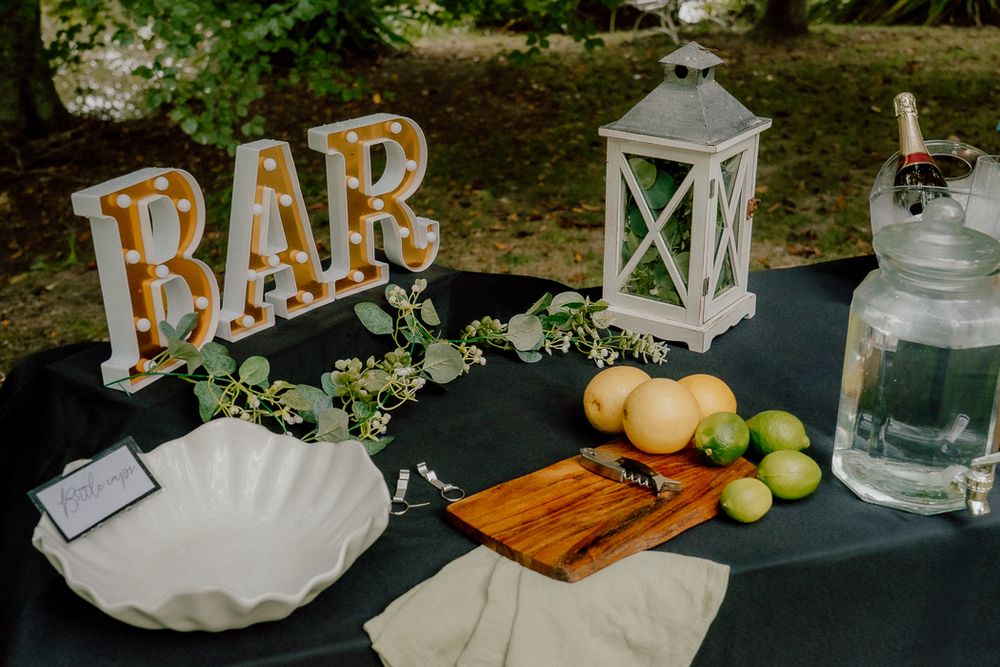 Outdoor wedding bar setup with marquee BAR letters, lantern, cutting board, limes, and glassware on a dark tablecloth.