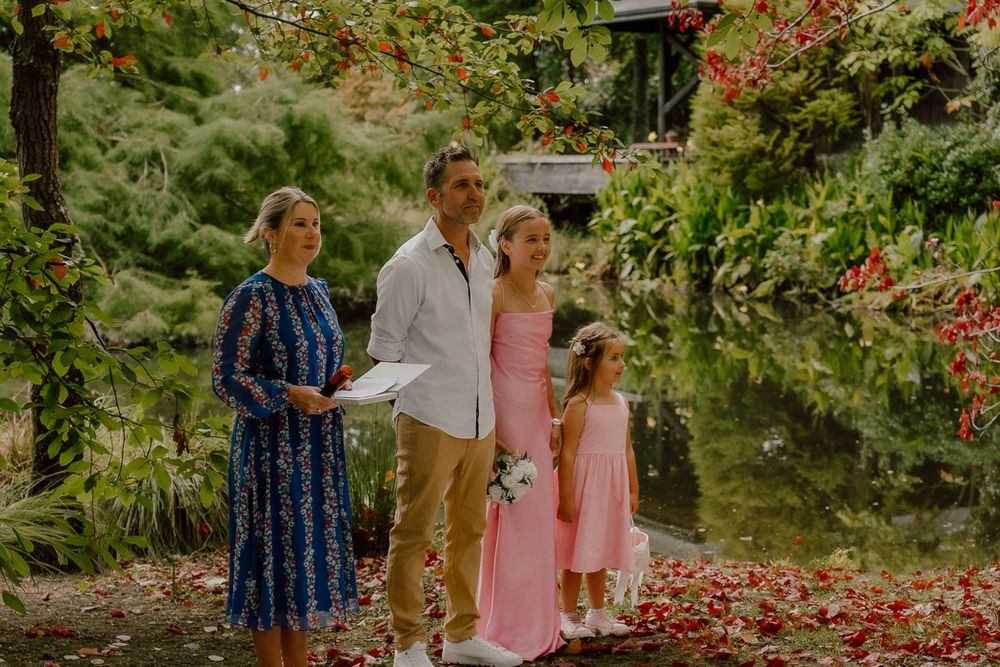 Outdoor wedding ceremony with couple, bridesmaid, and flower girl by a pond surrounded by lush greenery.