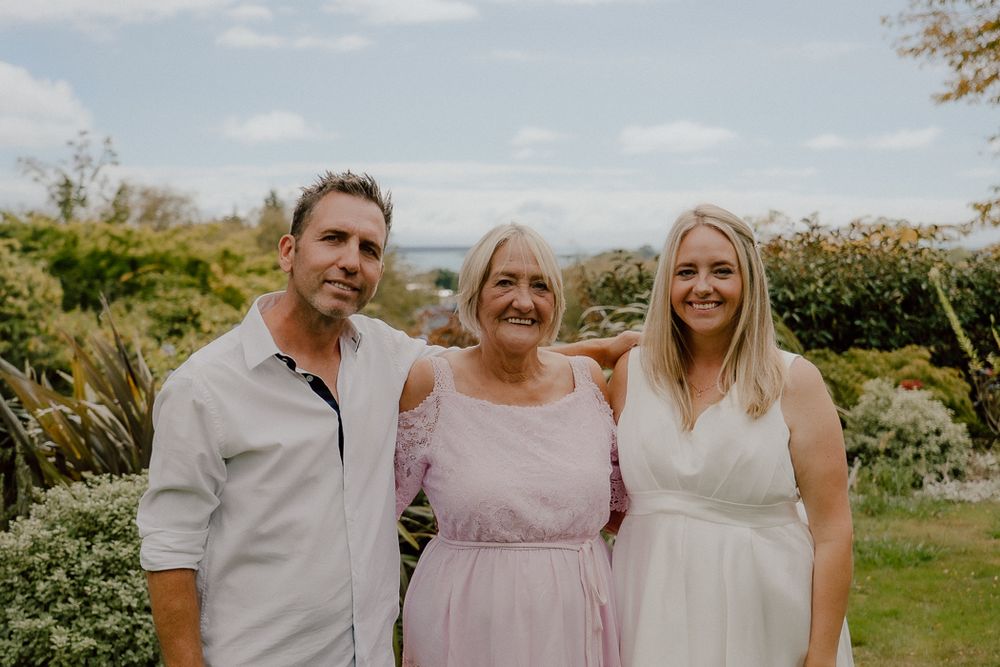 Three smiling people posing together in a lush garden, wearing casual and formal attire on a sunny day.