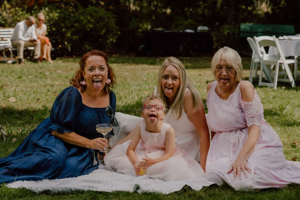 Four women laughing and sitting on grass at outdoor wedding, including flower girl in pink dress holding cocktail glass.