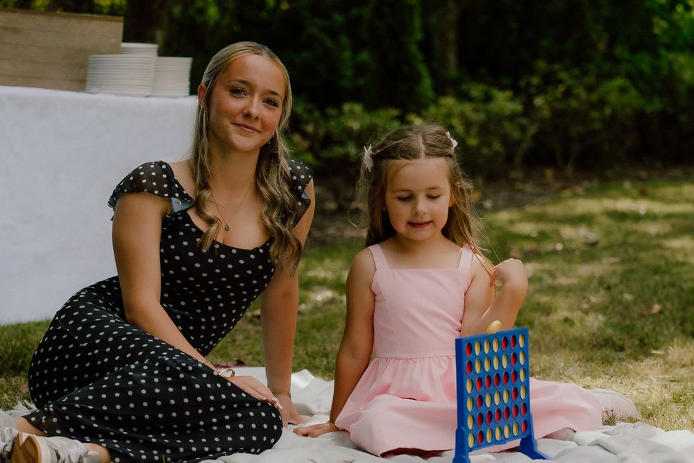 Two girls sitting on a blanket outdoors playing Connect Four board game in a garden setting.