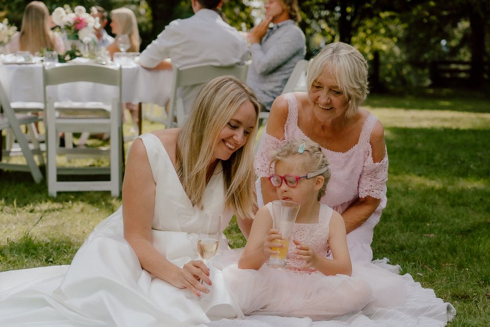 Bride in white gown sits on grass with young girl in pink dress and older woman at outdoor wedding reception.