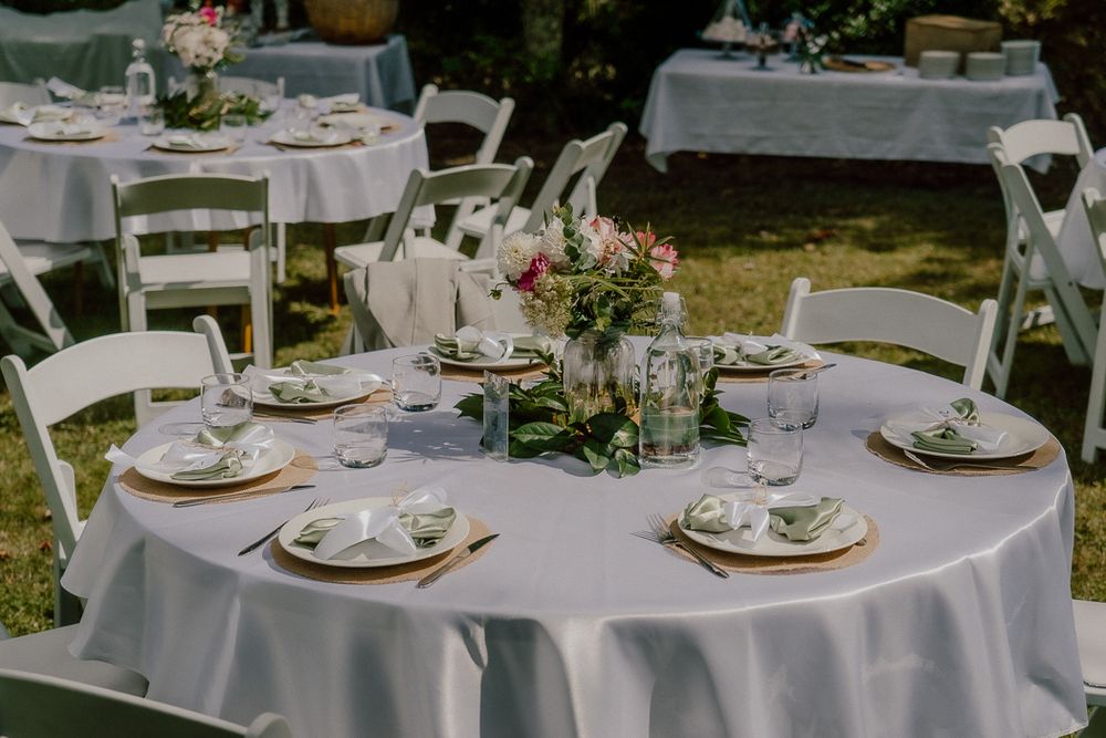 Elegant outdoor wedding reception tables with white linens, gold chargers, floral centerpieces, and white folding chairs on green grass.