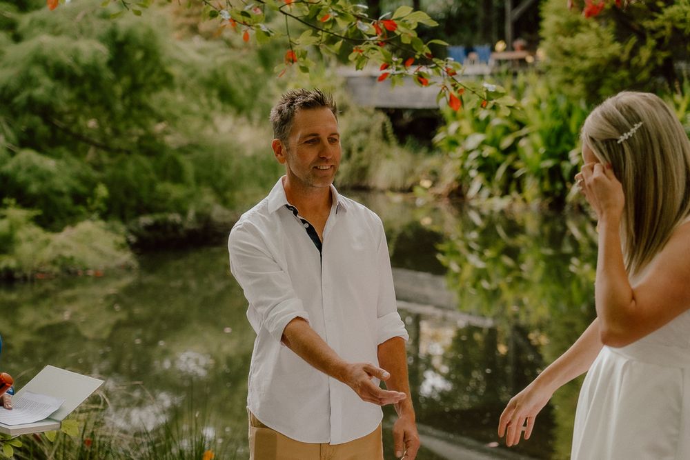 A groom in a white shirt holds hands with his bride during an outdoor wedding ceremony surrounded by lush greenery.