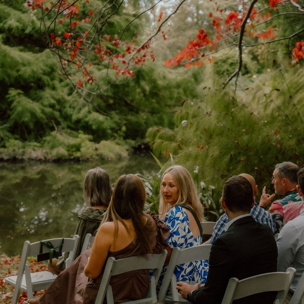 Guests seated in white chairs by a serene pond surrounded by lush greenery and vibrant red autumn foliage.