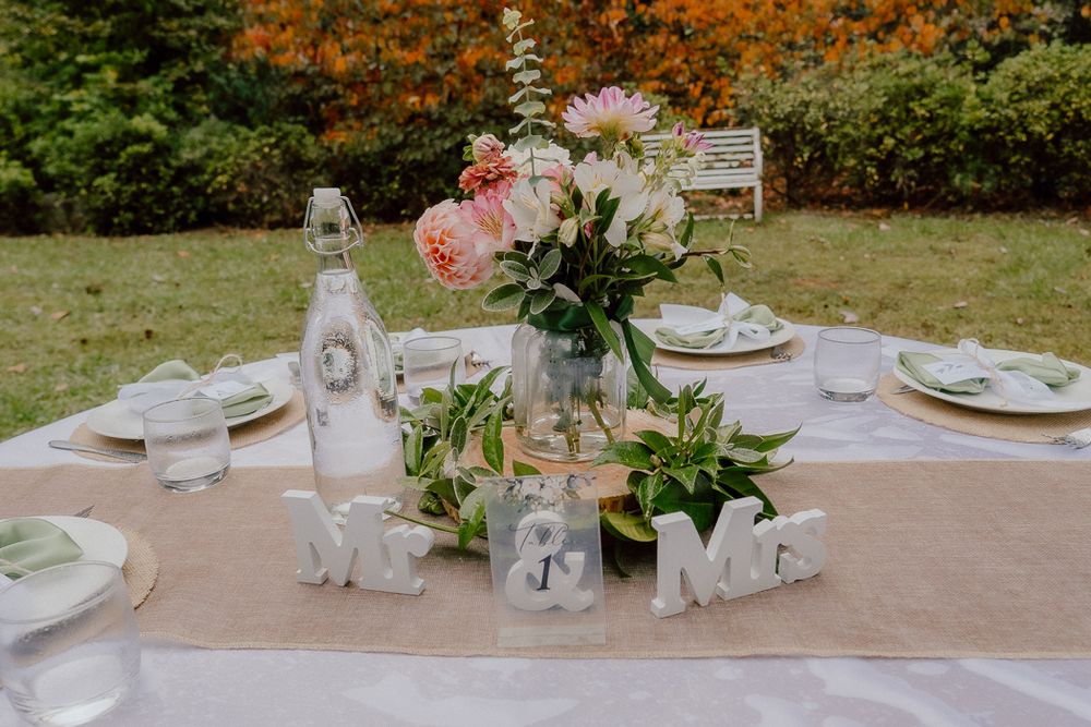Outdoor wedding reception table with Mr & Mrs signs, pink floral centerpiece, and elegant place settings on burlap runner.