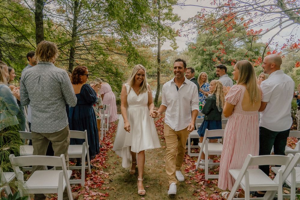 Bride and groom walking down outdoor wedding aisle lined with white chairs and red flower petals among autumn trees.