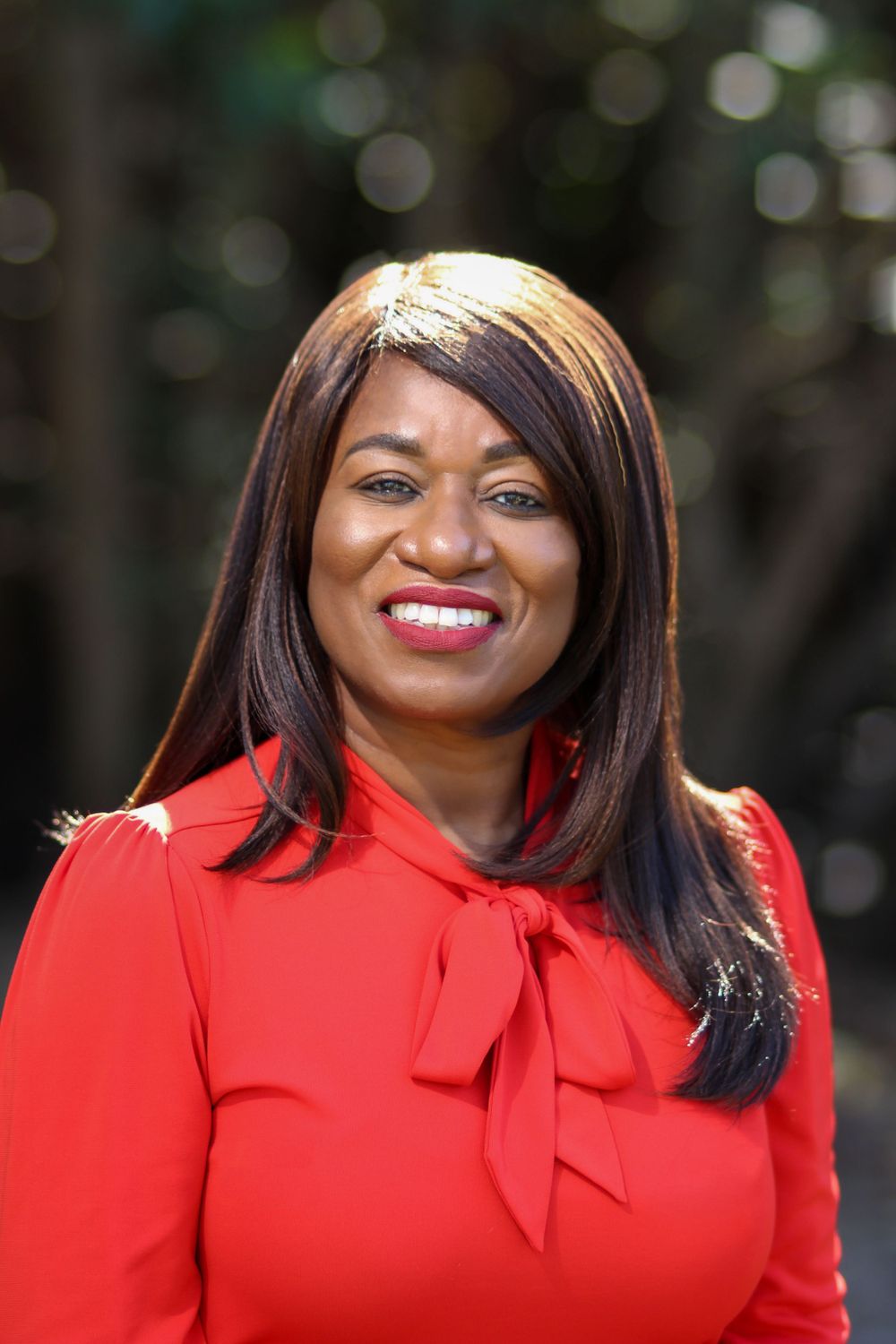 Professional portrait of person wearing vibrant red blouse with bow detail against blurred natural background.