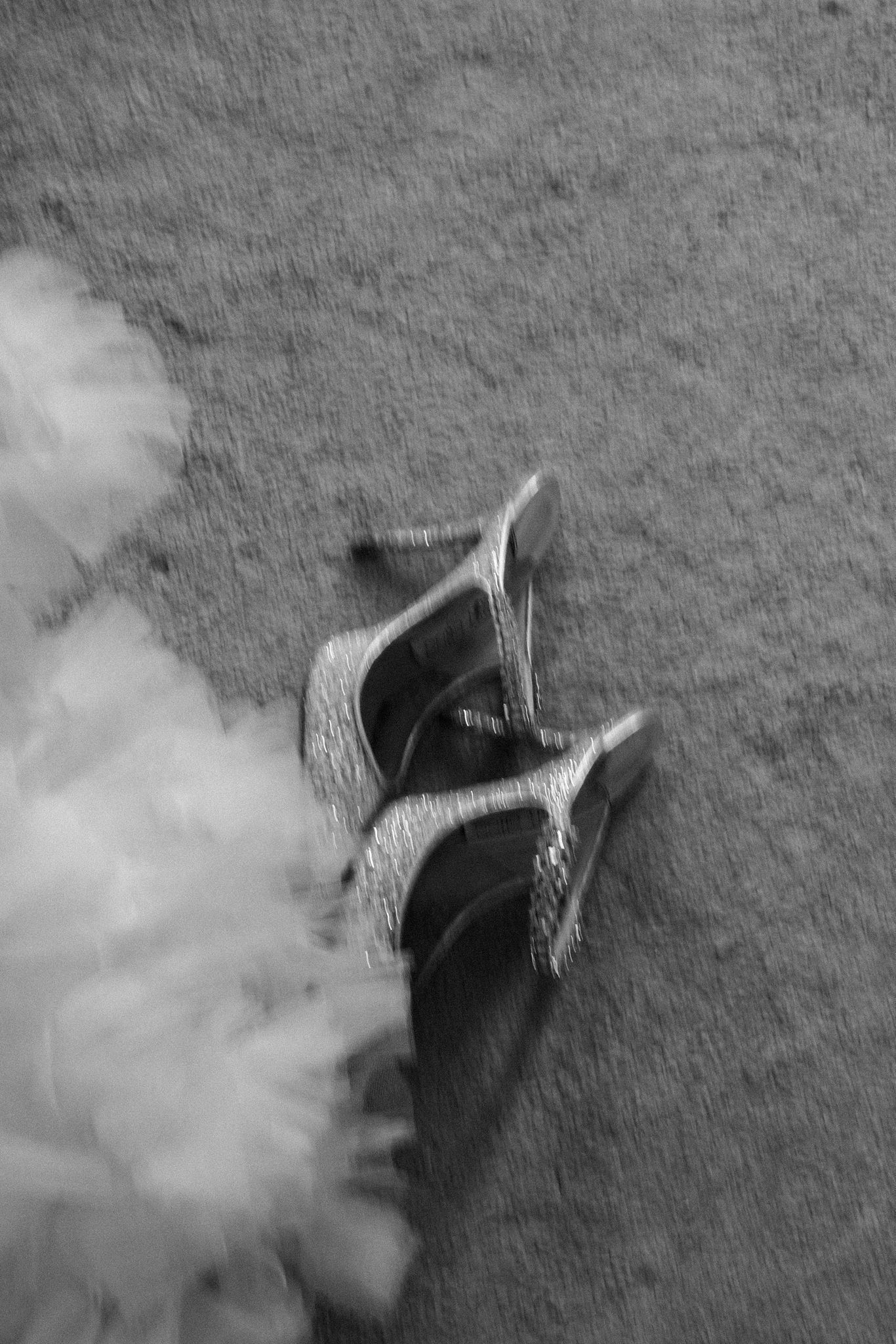 A pair of ornate silver high heel Jimmy Choo shoes on a textured gray surface with soft blurred elements in black and white.