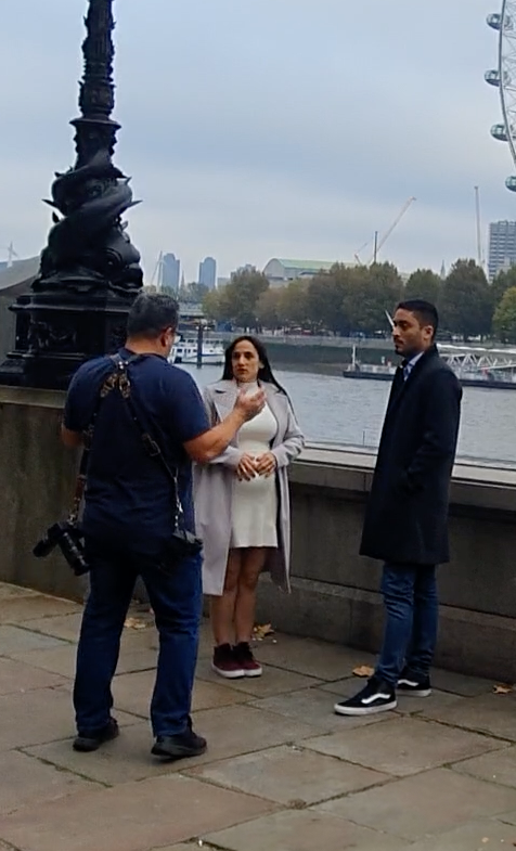 People being filmed or photographed along the Thames River in London with the London Eye visible.