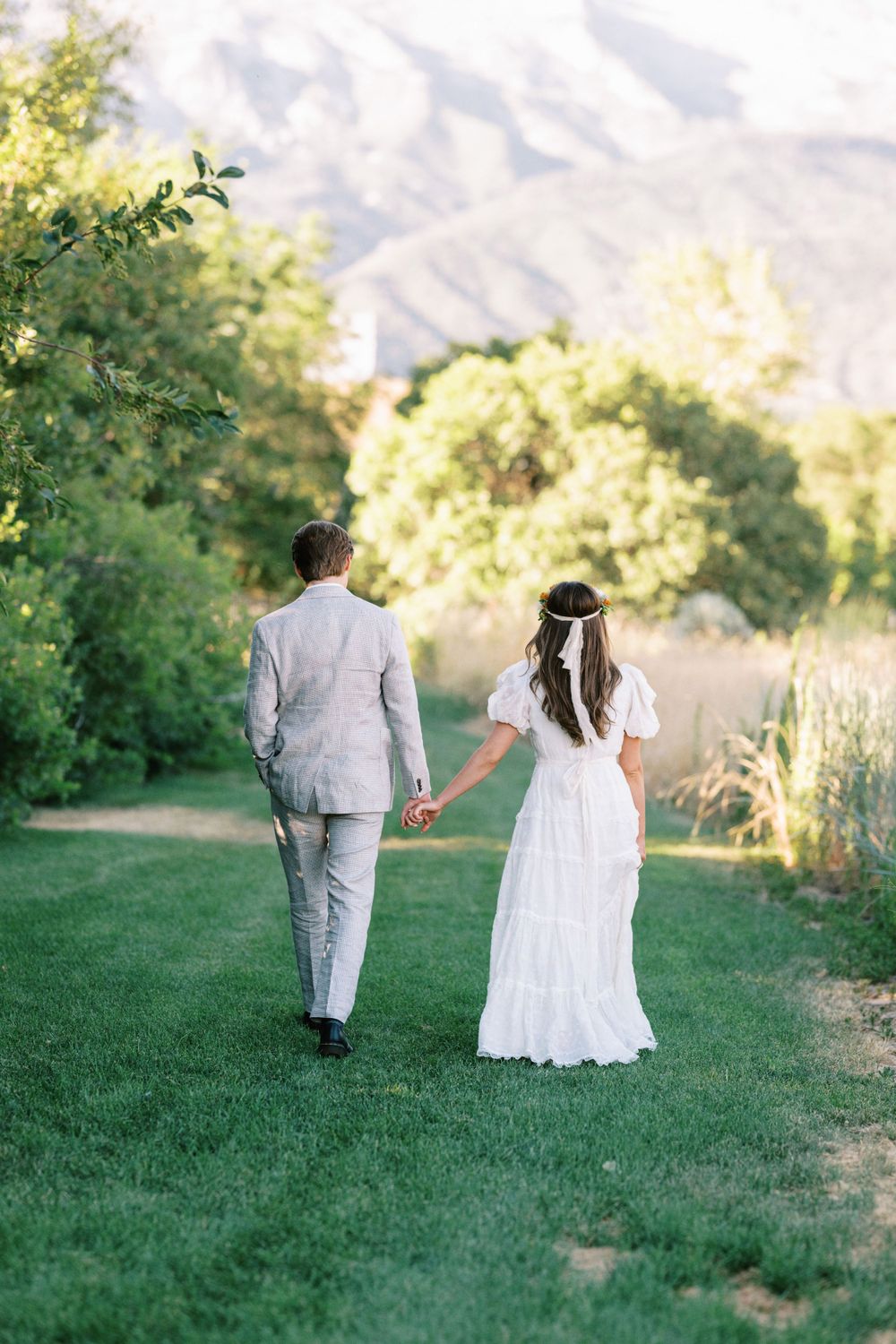 Wedding couple holding hands while walking on a lush green lawn with mountains and trees in the background.