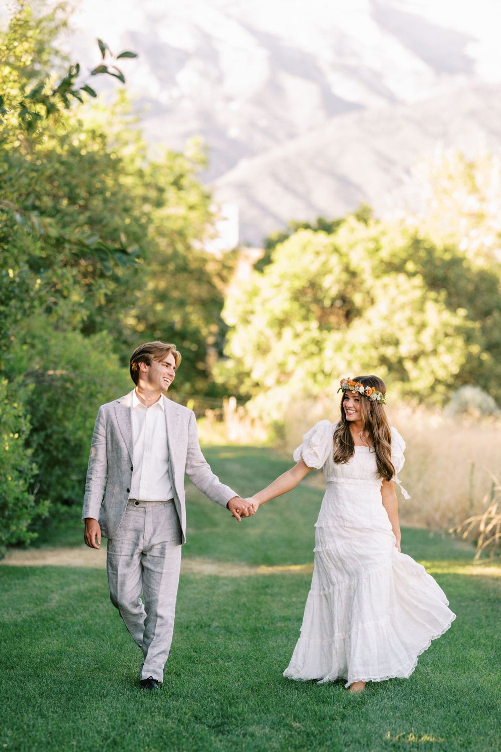 A couple in wedding attire walks hand in hand on a grassy lawn with mountain scenery and lush greenery in the background.