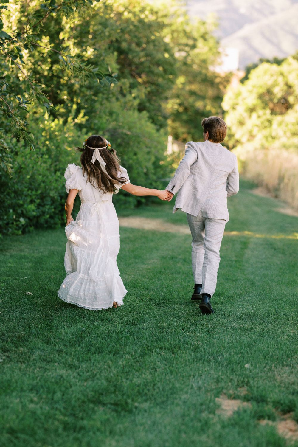 A couple in wedding attire runs playfully through a lush garden path on a sunny summer day.