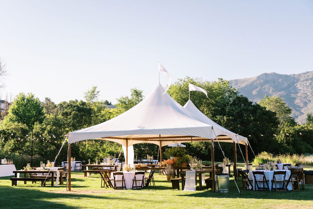 White wedding tent set up on a grassy lawn with round tables and chairs beneath mountains and trees in the background.