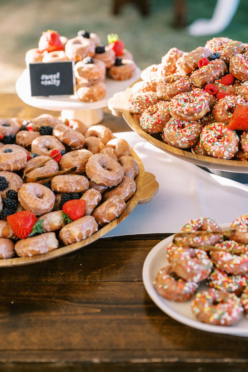 Assorted decorated donuts with sprinkles and fresh strawberries arranged on wooden platters at a dessert table.