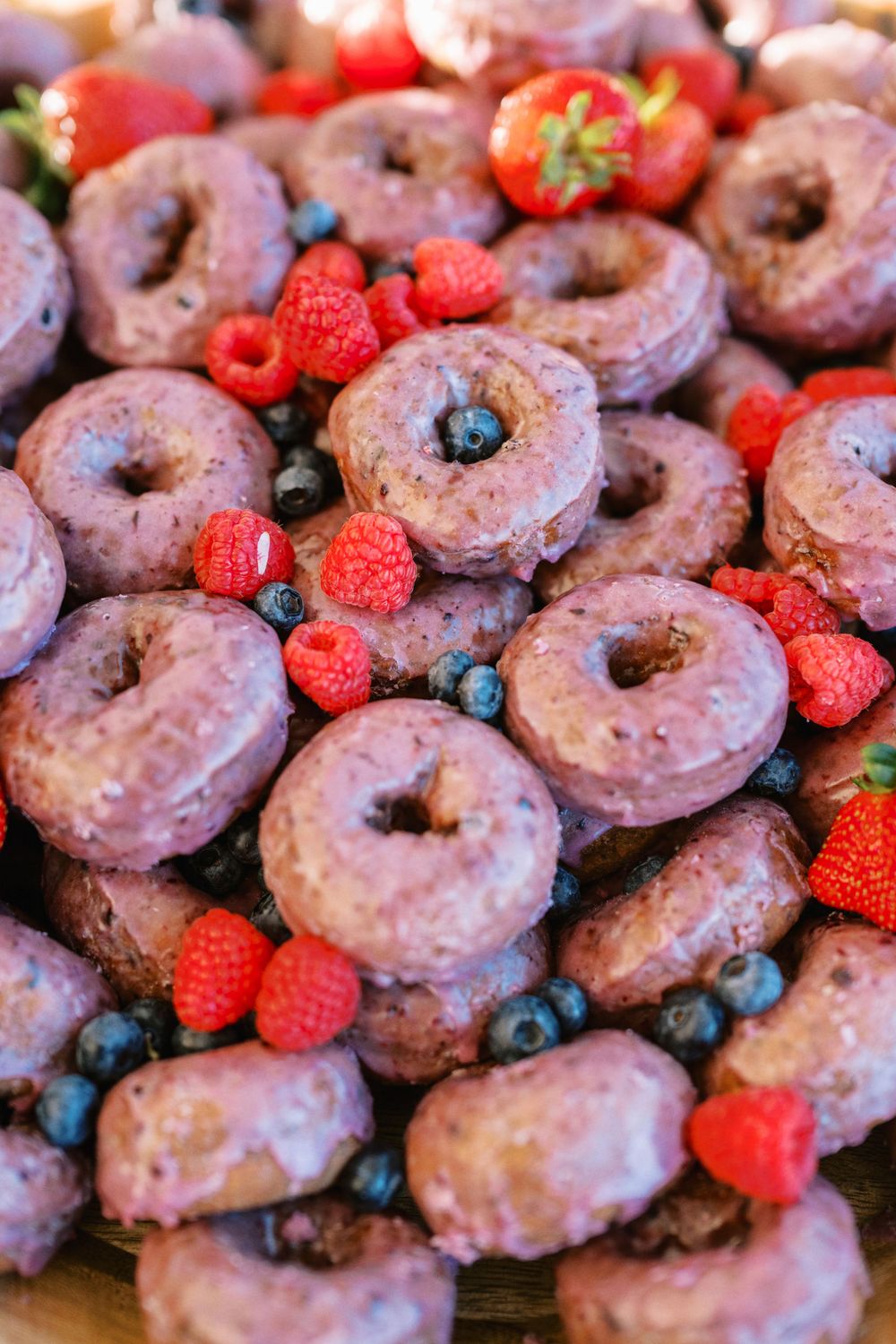 Chocolate glazed donuts arranged with fresh raspberries, blueberries and strawberries on a dark surface.