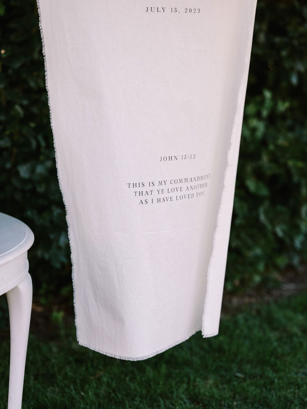 A white fabric wedding banner with date and text displayed against a green hedge backdrop.
