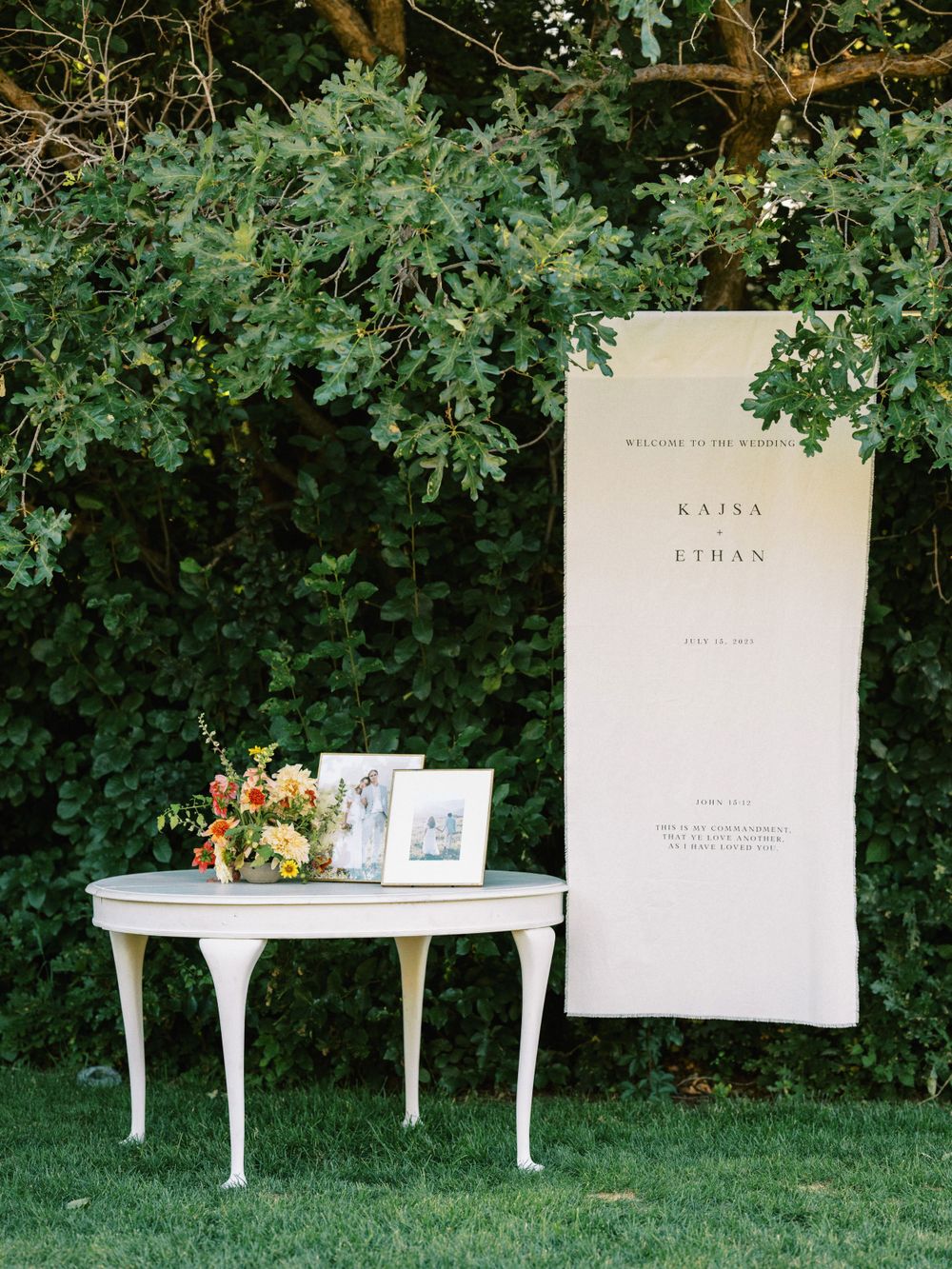Elegant white wedding display with side table and banner against lush greenery backdrop.