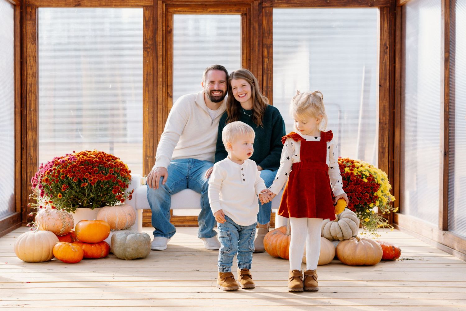 A family poses with autumn decorations including pumpkins and red mums on a sunlit porch.