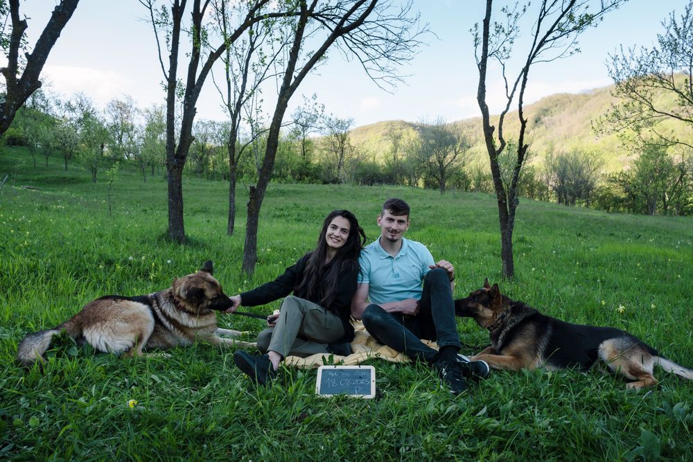 A couple relaxes with their German Shepherd on a grassy hillside with trees in the background.