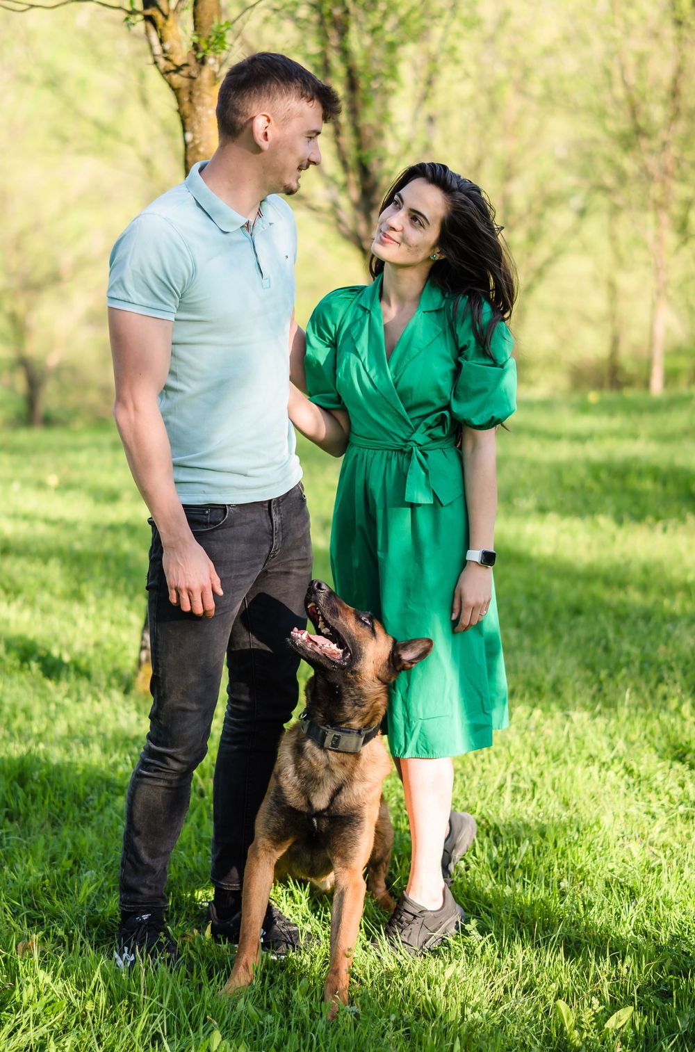 A couple poses with their German Shepherd dog in a sunlit grassy field.