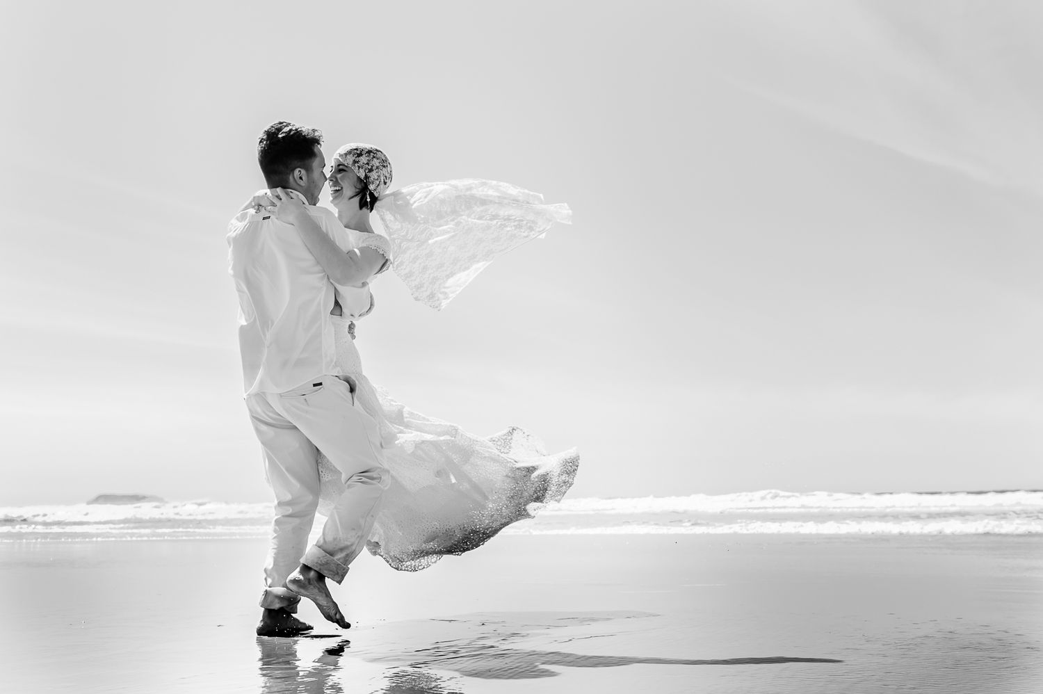 A couple in white attire embracing on a beach with flowing fabric creating an ethereal effect in black and white photography.
