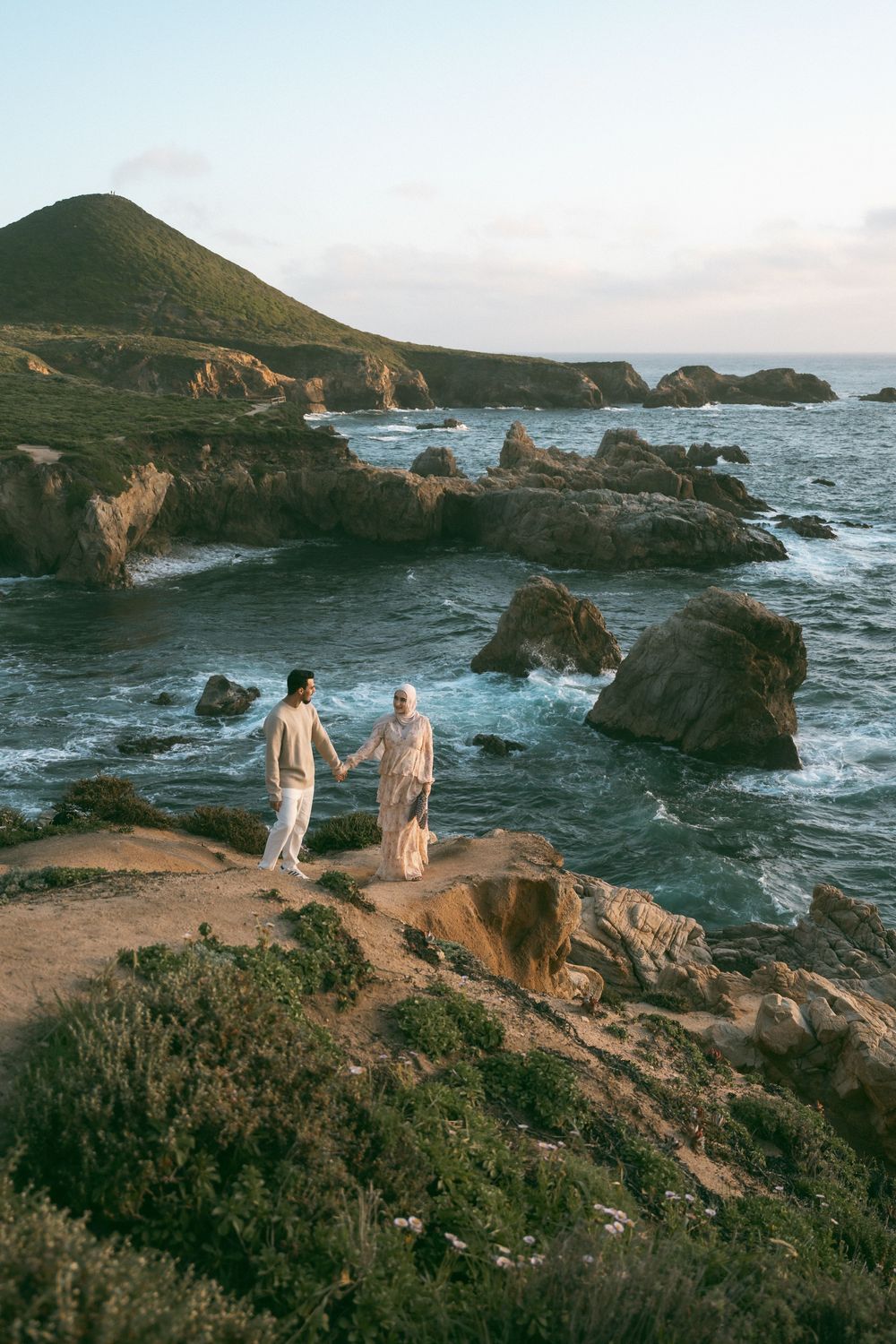 Couple holding hands on rocky coastal cliffs with ocean waves and green hills at sunset.