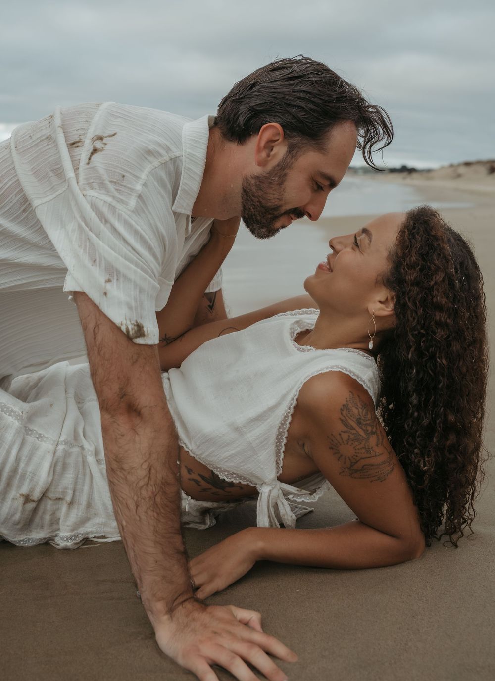 Romantic couple in white outfits embrace intimately on a sandy beach during an overcast day.