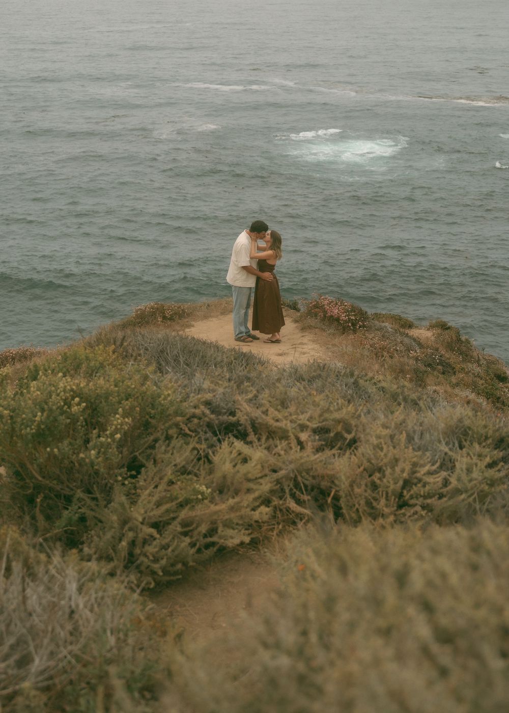 Couple embracing on a rocky coastal cliff overlooking the ocean, surrounded by wild shrubs and misty sea views.