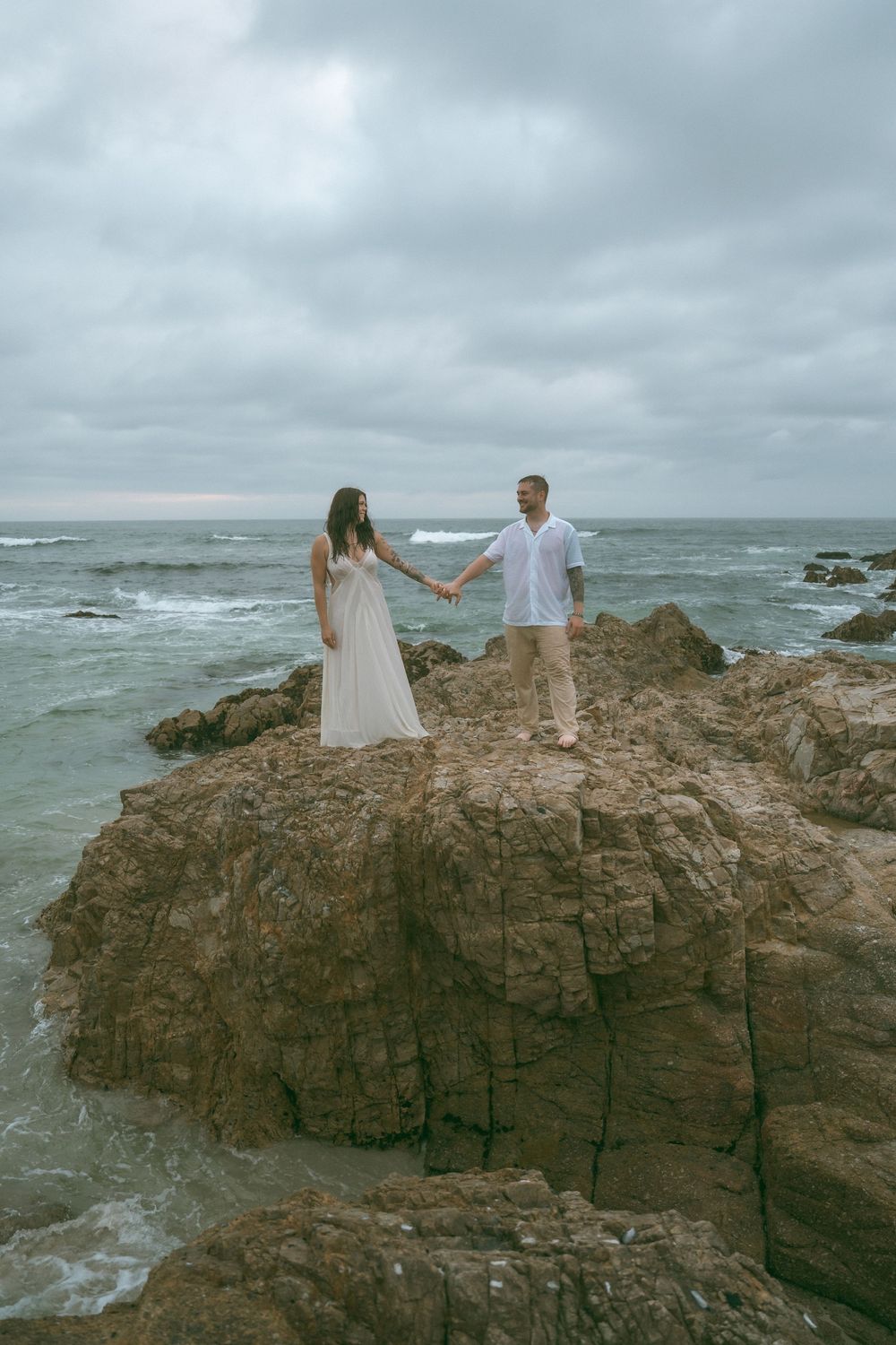Couple holding hands on rocky coastal cliffs with ocean waves and cloudy sky, wearing white dress and casual attire.