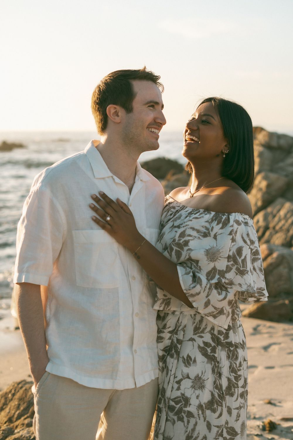Smiling interracial couple embracing on a rocky beach at golden hour, woman in floral off-shoulder dress.