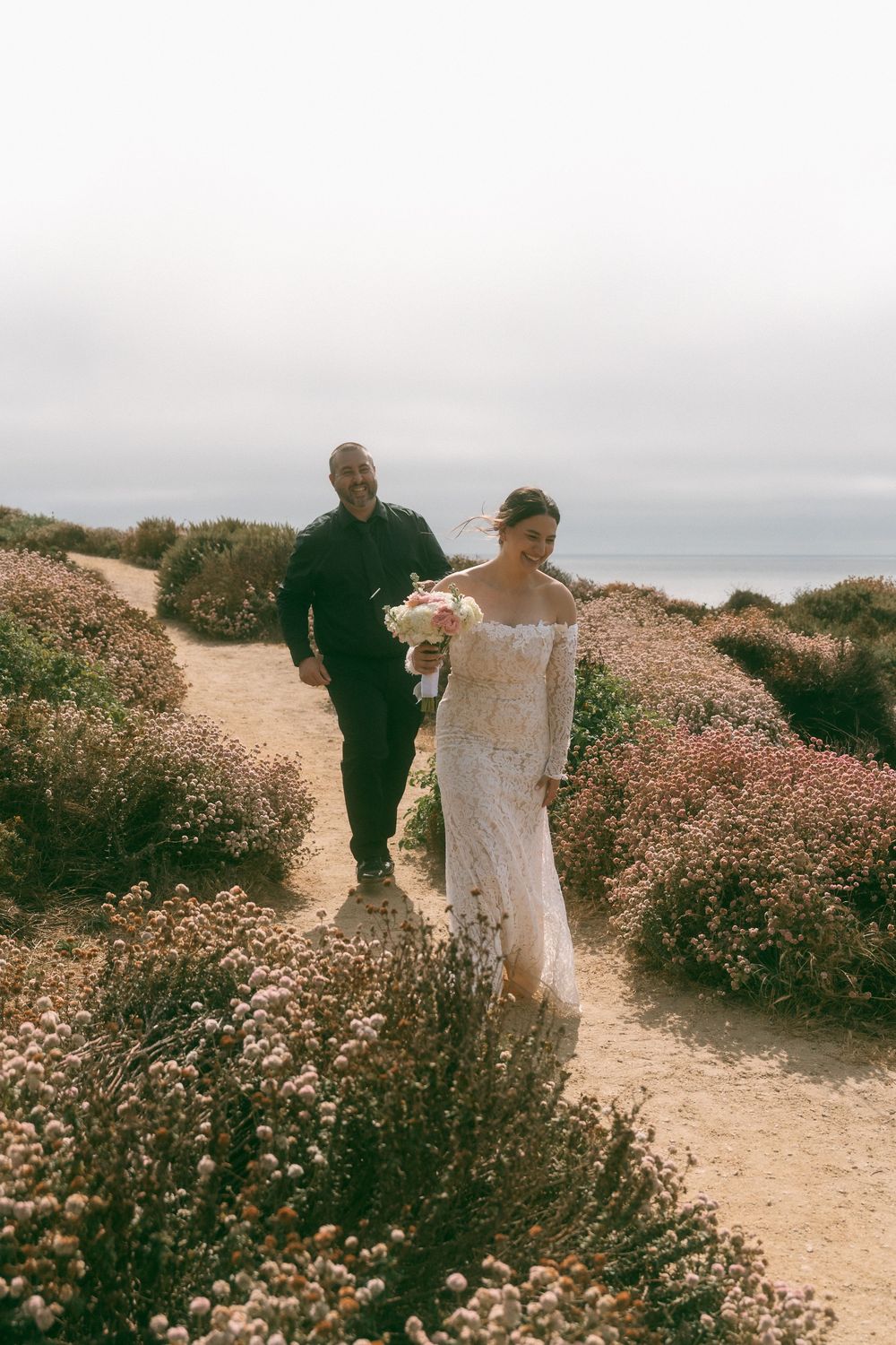 Bride in lace gown holding bouquet walks coastal wildflower path with groom in black, ocean backdrop.