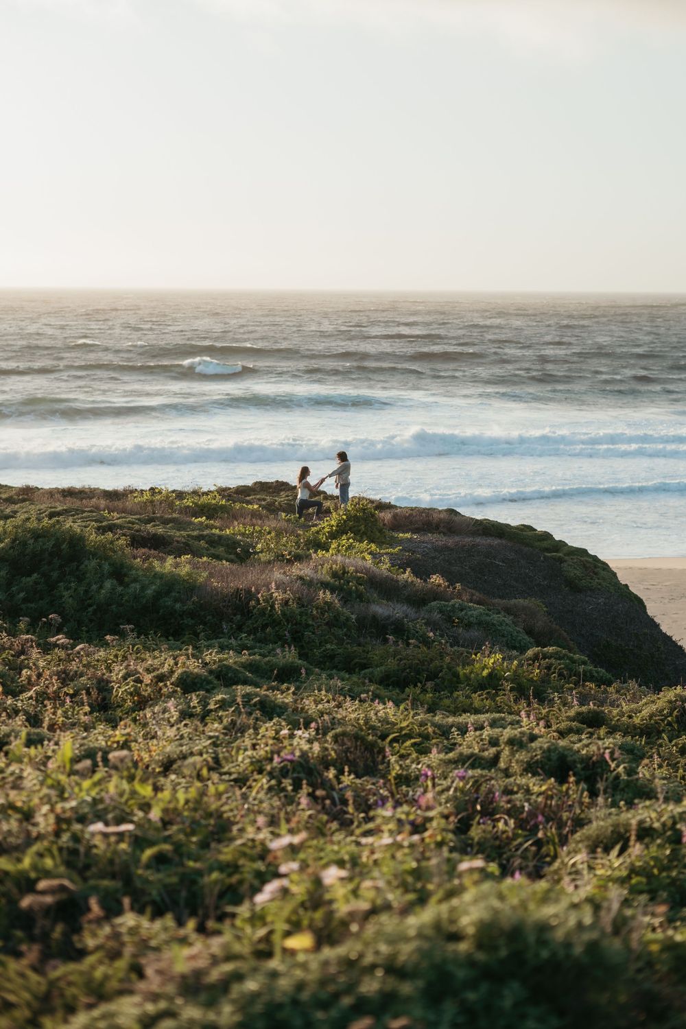 A scenic coastal landscape with people standing on a cliff overlooking the ocean waves and vegetation at sunset.
