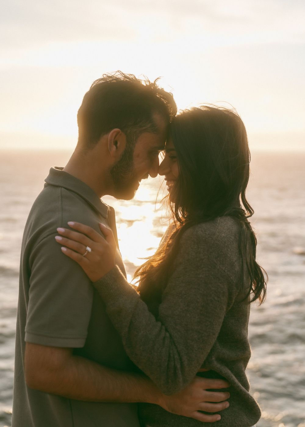 Romantic couple embracing at sunset by the ocean, sharing an intimate moment with golden light behind them.