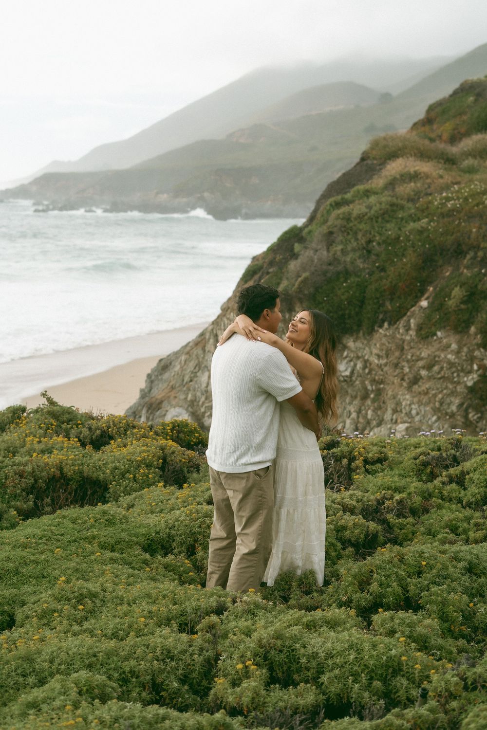 Couple embracing on a coastal bluff with misty mountains and ocean waves in the background.