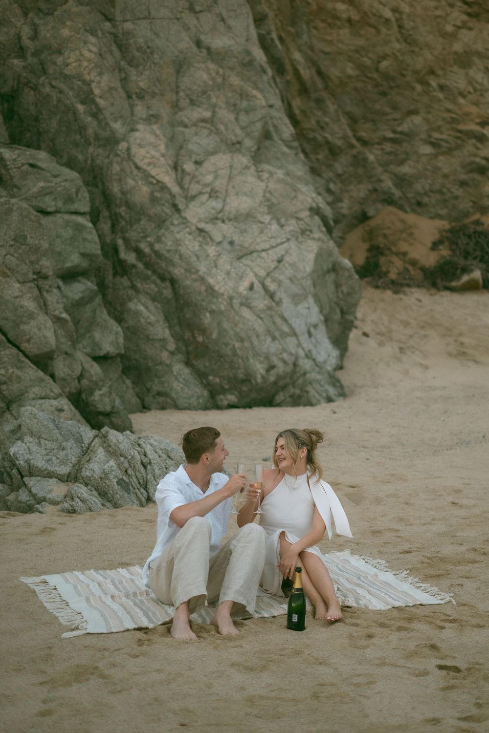 Couple toasting champagne glasses on a beach blanket with rocky cliffs behind them during a romantic seaside picnic.
