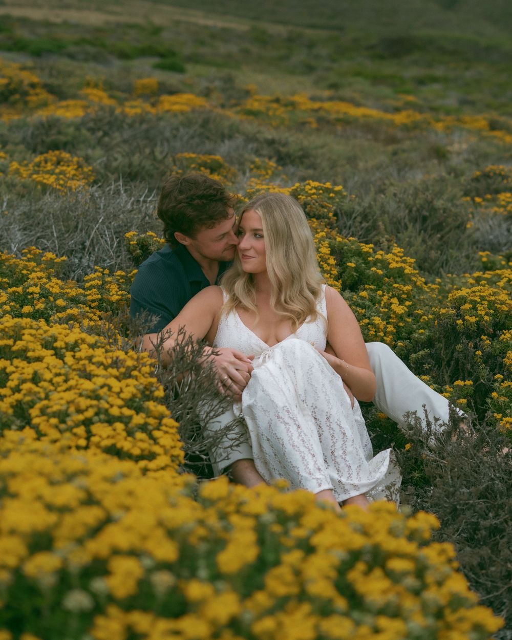 Couple sitting romantically in a field of yellow wildflowers, woman in white dress, man embracing her from behind.