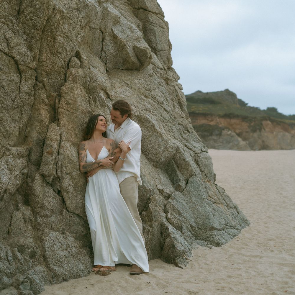 Couple in white attire leaning against rocky cliff on sandy beach during romantic outdoor photo session.