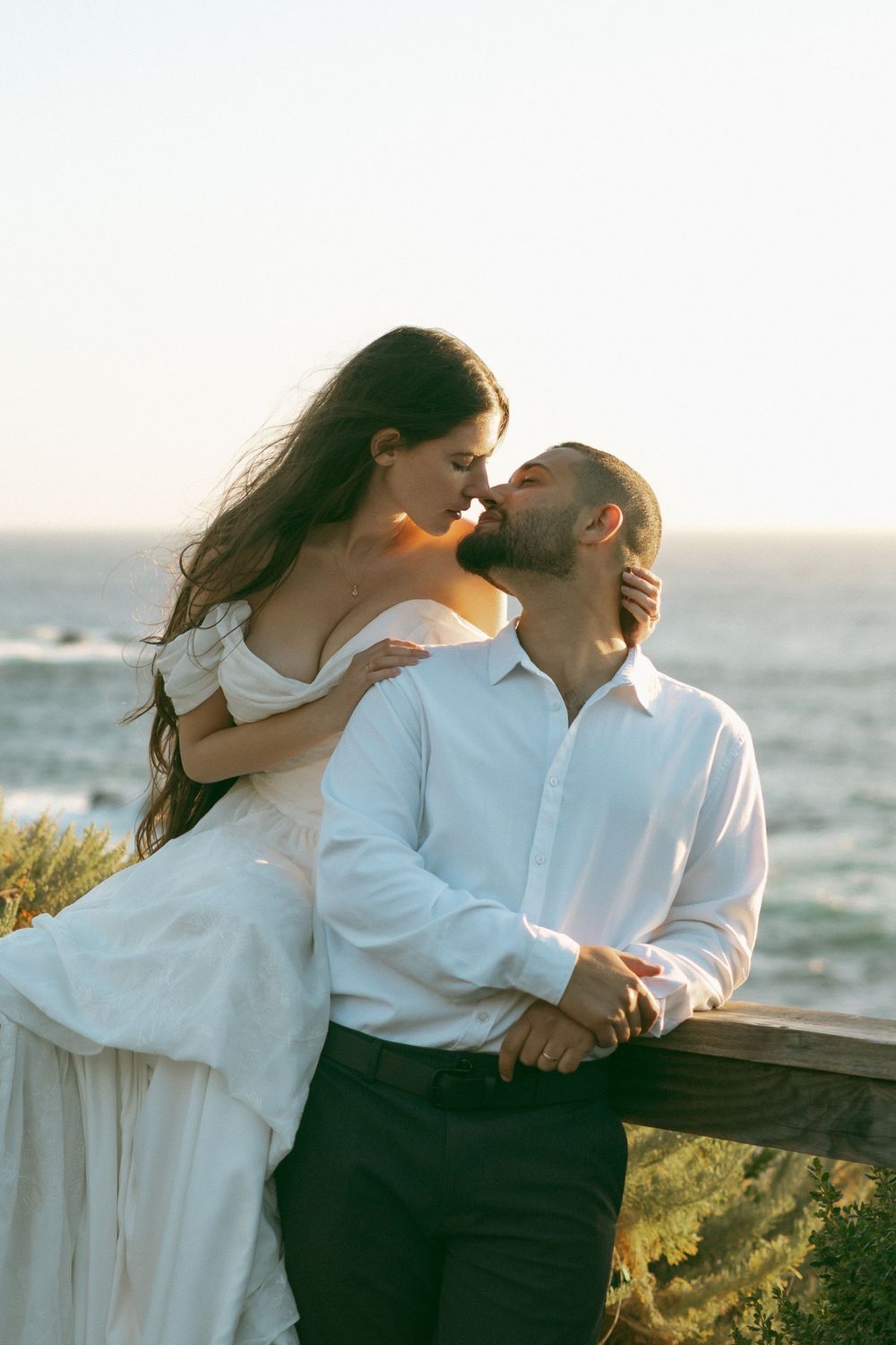 A couple in white attire share a romantic kiss at sunset by the ocean, with the bride seated on a wooden railing.