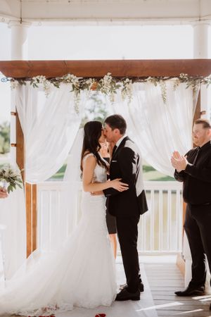Newlyweds share their first kiss under white draped wedding arch decorated with delicate flowers during outdoor ceremony.